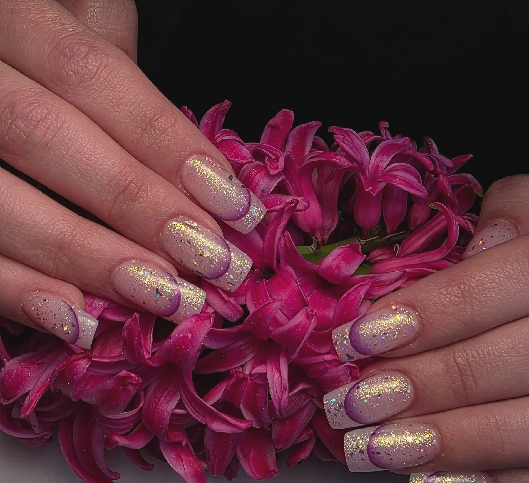 Lange vierkante nagels met holografische glitter en een paarse french-rand, op roze hyacinten tegen een zwarte achtergrond